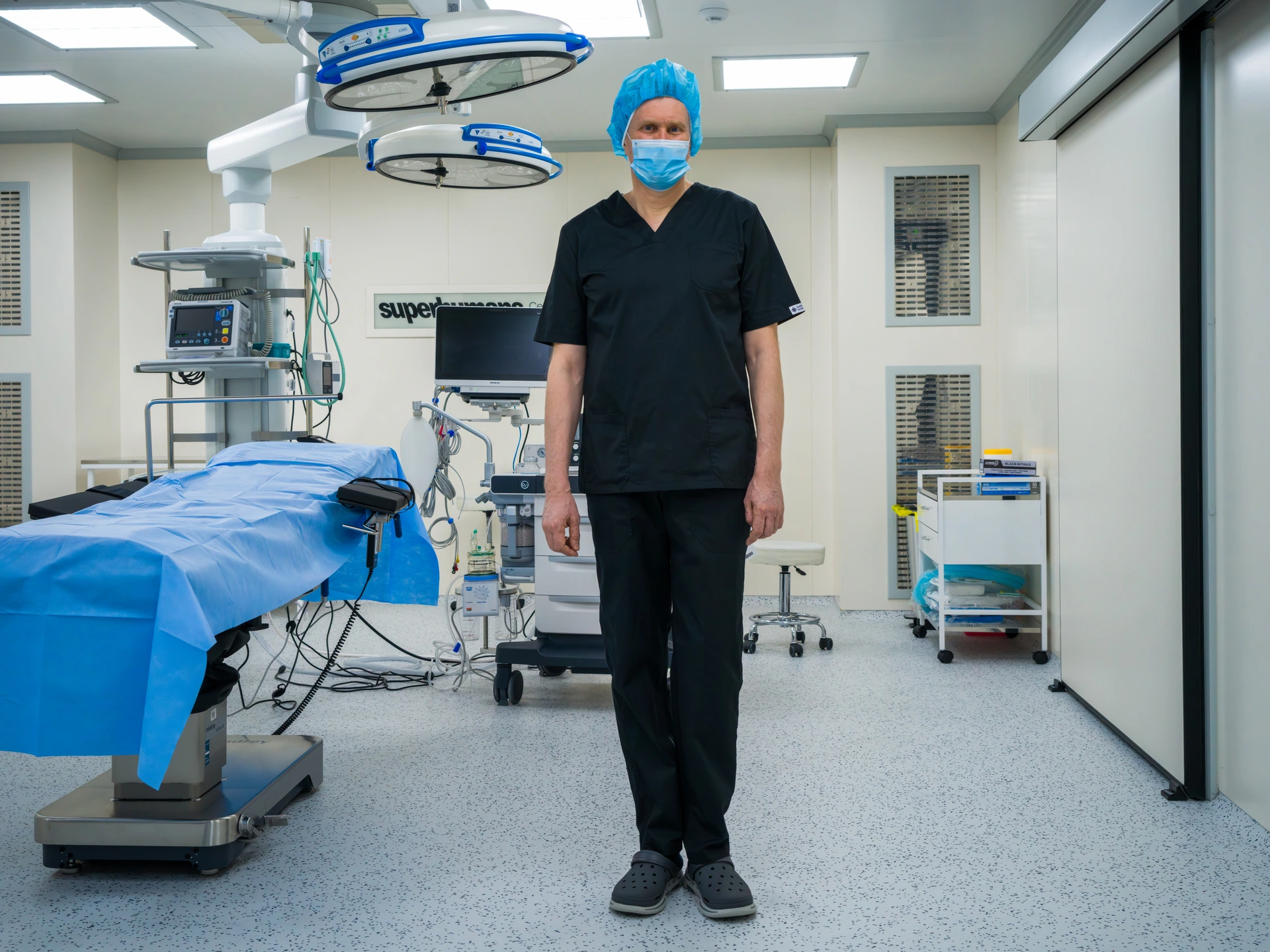 A man stands in a brightly lit operating theatre, looking straight at the camera, with an operating table to his right and surgical equipment and a light stand in the background. The man is wearing a light blue surgical cap and mask, a dark blue hospital uniform, and dark blue plastic slip-on shoes. The man is Andrii Vilensky, the Medical Director of the Superhumans Centre, a clinic in the western Ukrainian city of Lviv that provides expert prosthesis and rehabilitation treatment to Ukrainians injured in Russia's full-scale war on the country.