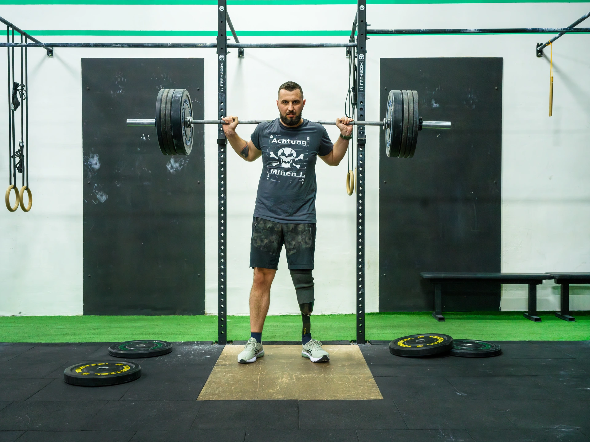 In a gymnasium, a man stands on padded flooring holding a weight lifting bar over his shoulders with several weights connected on both sides of the bar. He is wearing a t-shirt with a white skull and crossbones between the German words "Achtung Minen" printed on the front. He also has a small tattoo on his right forearm. He is wearing shorts, revealing a prosthetic left leg. Several weights are scattered on the floor around him. He is sporting a small dark beard with buzz-cut dark hair, and is staring intently at the camera with a look of defiance. This is Ihor Bezkaravainyi, the 34 year-old Deputy Minister of Economy of Ukraine and Invictus Games athlete, who lost his leg during fighting in Donetsk Oblast in 2015.