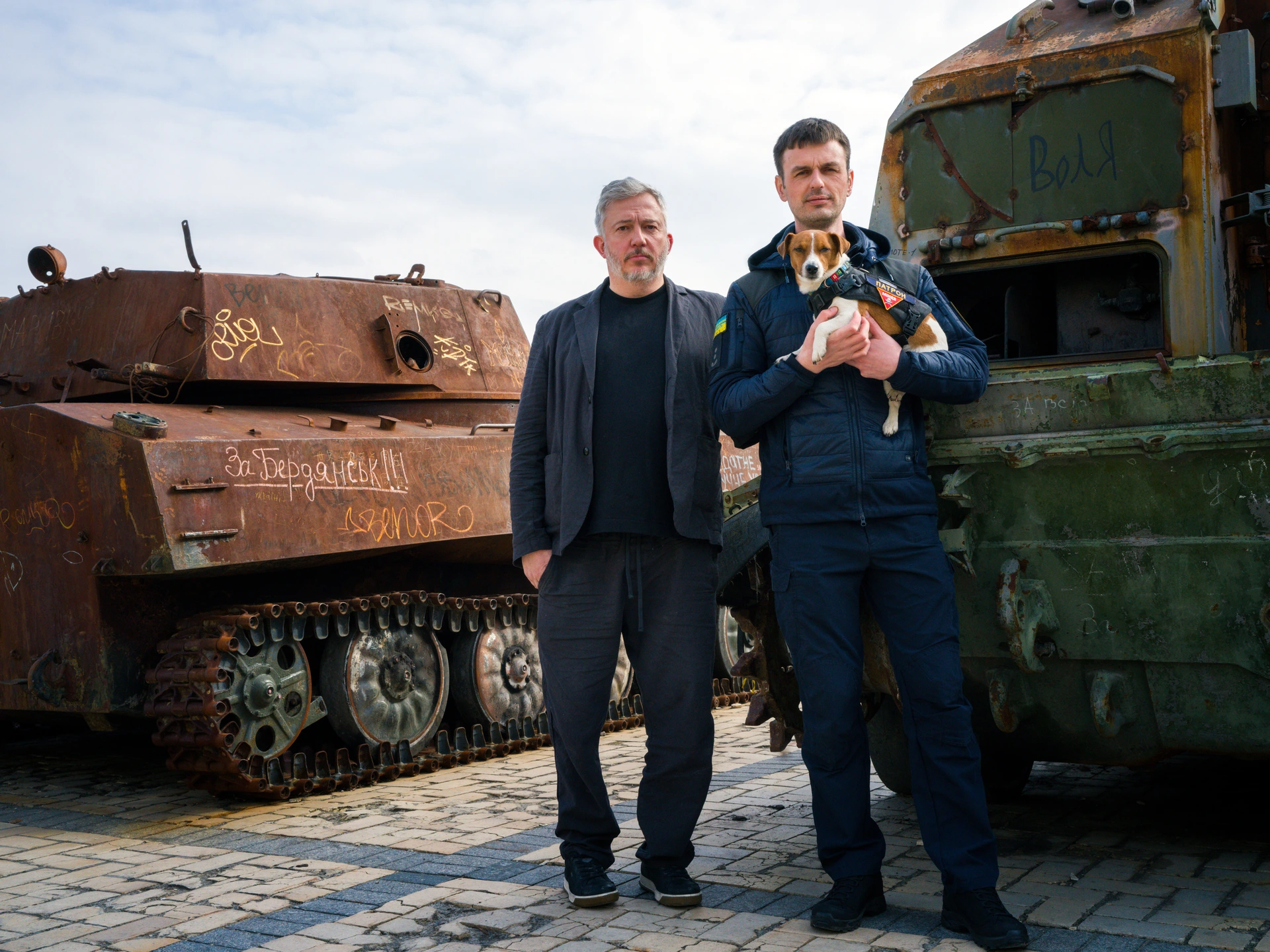 Two men, one holding a dog, stand outside in front of a rusty, abandoned tank with graffiti etched into the metal bodywork and an abandoned, rusted, green military vehicle with missing window glass. The man on the left, photographer Giles Duley, has short grey hair and a goatee, and is wearing a navy blue jacket and black t-shirt. Next to him is sapper Mykhailo Iliev, a younger man with short, brown hair, who is wearing a dark blue tracksuit with a flag of the Ukraine stitched to his right arm. Mykhailo is holding a small brown and white Jack Russell sniffer dog named Patron, who is wearing a small bulletproof jacket over his fur.