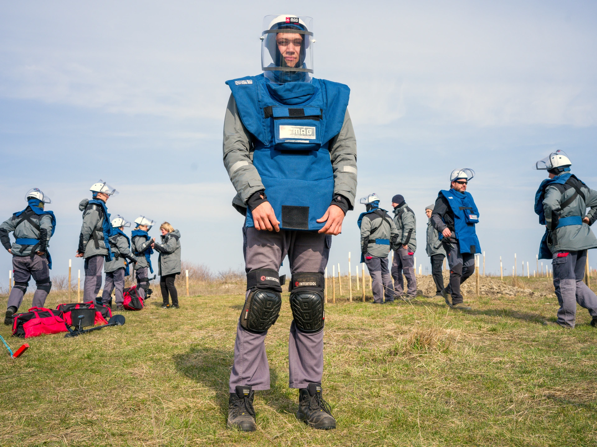 A man stands in a field with short grass, and a blue sky covered with wispy clouds. He is wearing black, ankle-high boots, mauve trousers, black kneepads, a grey jacket with visibility stripes on its sleeves, and a blue protective vest. He is wearing a white helmet over a black hood, and a transparent plastic visor covers his face. His hands are gripping the bottom of his blue protective vest, at the front, on either side. Behind him on both sides are two groups of similarly dressed people, who are standing next to an area marked off by wooden posts painted white on their tops. They all face away from the camera, looking ahead in the same direction. The man is Oleksander Kuchen, a deminer with Mines Advisory Group, a non-governmental organization that assists people affected by landmines, unexploded ordnance, and small arms and light weapons. He is pictured in Taborivka, a village in Mykolaiv Oblast, in the south of Ukraine.