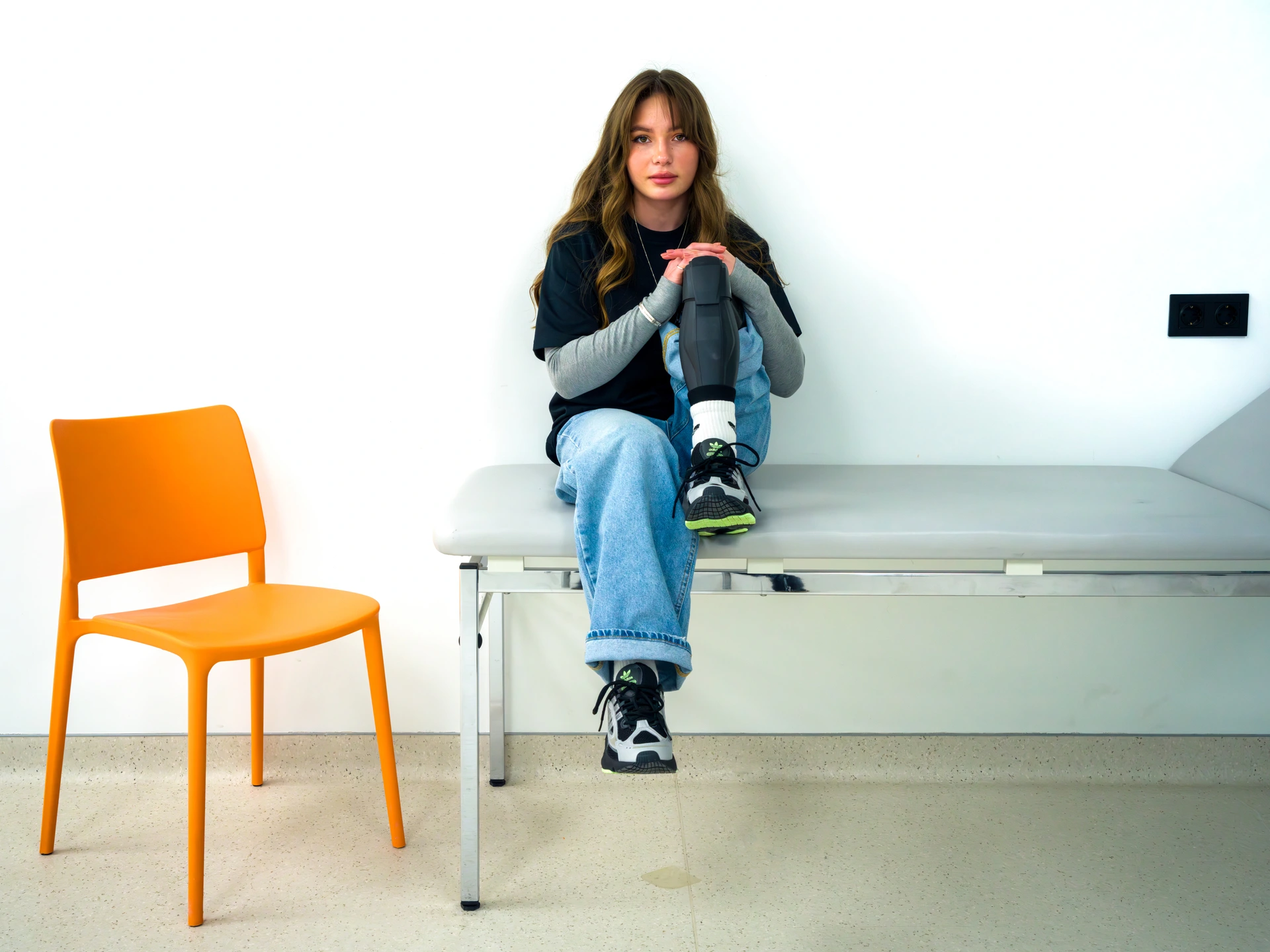 A girl, staring at the camera with a fixed expression, sits on a grey examination bed in a bare, white-walled room, with an orange plastic chair to her right. Her clasped hands are resting on the knee of her left leg, which is prosthetic and made of black plastic, its foot resting on the examination bed, while her right leg dangles in the air above the bed. She has long brown hair and is about 20 years old. She is wearing black and green trainers on both feet, baggy blue denim jeans, and a black t-shirt over a grey, long-sleeved sweatshirt. This is Rusya Danilkina, Advocate and first Contact of the Superhumans Centre in the city of Lviv in western Ukraine.