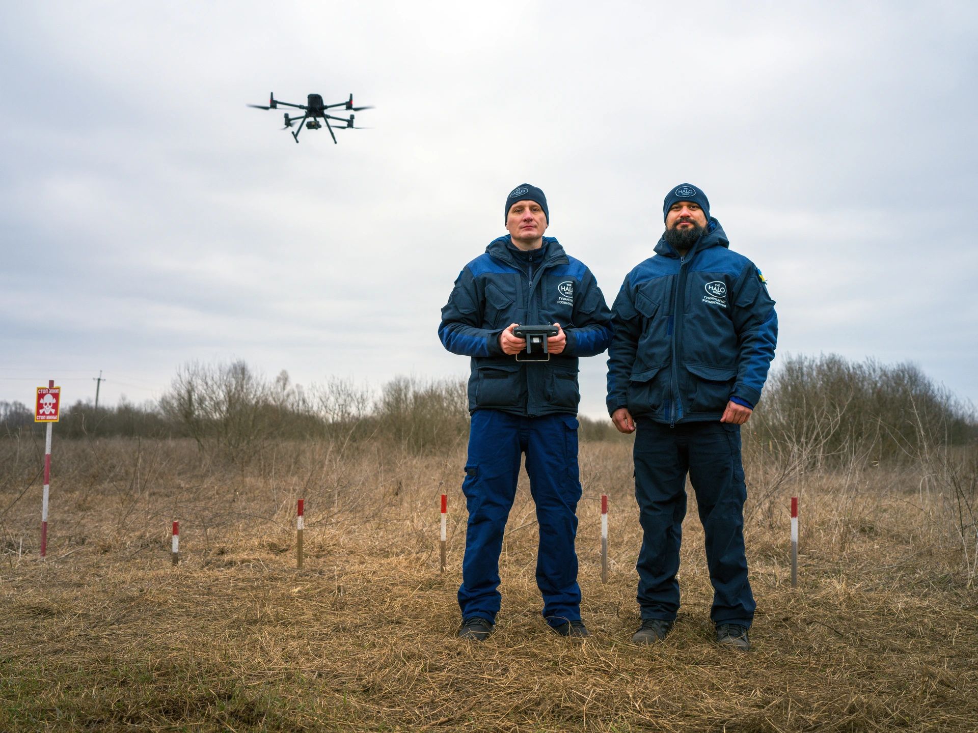 Two men are standing outside, on a patch of brown scrubland with bushes in the background, under a dull, cloudy sky. Behind them is an area marked off with red and white painted posts, and a white and red "Danger: Mines" sign with a skull and crossbones. To the right of the men a small, quadcopter drone hovers in the sky, just over head-height. Both men are wearing matching, blue waterproof trousers and jackets, and dark blue woolly hats. The man on the left, Serhii Pasholok, is clean-shaven and holding a drone control unit. Next to him is Selim Granqvist-Ahmed, who has a black beard. Granqvist-Ahmed, who is 38, is a Research and Development Manager of the HALO Trust mine-clearing organization.