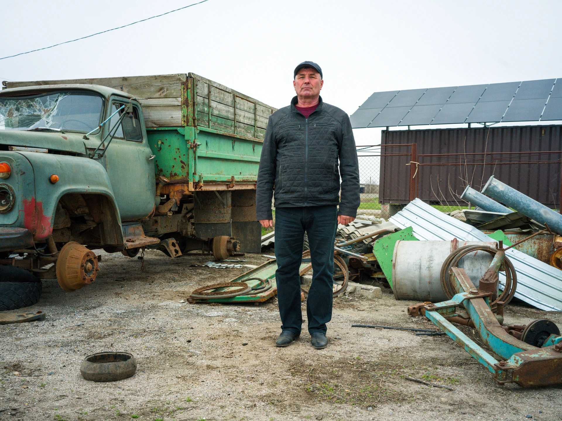 A middle-aged man stands looking at the camera. Behind him on the right, there's a green broken-down truck with both wheels missing on one side, revealing rusty axles. The windscreen is shattered and the front left headlight has been smashed off. To his left, rusty scrap pieces of metal and a metal barrel are scattered on the ground in an untidy pile, and behind them is a shelter with solar panels on its roof. The man is wearing a black jacket and cap, stands alone, with his arms by his side, and stares with pressed lips and a fixed look, squinting his eyes at the camera. He is Serhii Tsvetkov, a 58-year-old farmer from the village of Kalynivka, in Mykolaiv Oblast in the south of Ukraine.