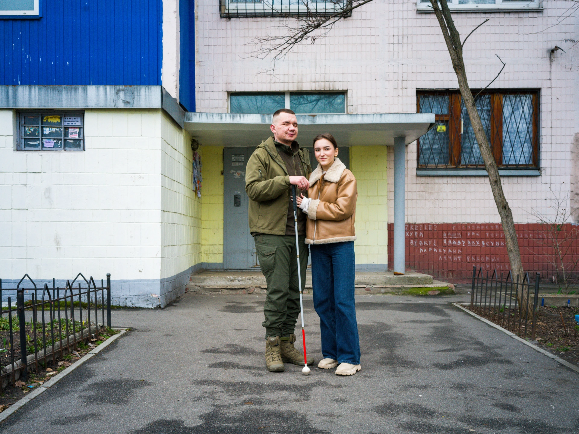 A man and a woman stand outside the entryway to a light-pink-tiled apartment building with brown-framed windows. The man, on the left, has short dark hair, closely cropped at the sides, and is wearing a khaki jacket with dark green combat trousers. He is holding a white cane and is facing away from the camera, with his eyes closed. He is Vladyslav Yeshchenko, the founder of Let's See The Victory charity which aids people who have lost their vision due to being injured in the war.The woman standing to the Vladyslav’s left is looking at the camera. She has dark hair tied in a ponytail, and is wearing a tan-coloured sheepskin jacket, baggy jeans, and beige shoes. She has her hand pressed on the man's abdomen, and is called Valeria Yeshchenko. She is the wife and co-worker of Vladyslav.