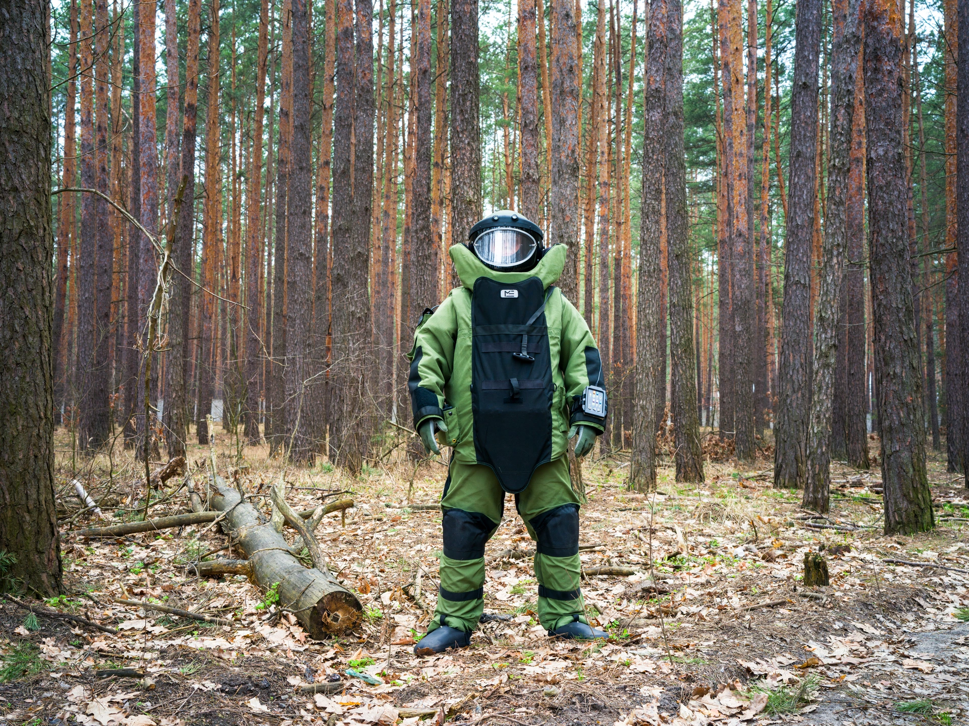 In the middle of some woods, an anonymous bomb technician for the State Emergency Service of Ukraine stands alone. Tall pine trees with brown and orange bark can be seen behind the technician, and the ground is covered in brown, dead leaves. To the right, a lone tree trunk lies on the ground. The technician wears a bulky, green and black body hazard suit with green gloves and a helmet covering their face. The light reflects off the transparent visor obscuring the person's facial features. The technician has an open stance, with their feet splayed.